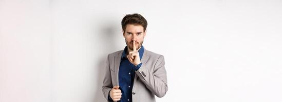Angry businessman showing taboo sign, shushing and tell to keep quiet, need silence, standing with finger on lips and looking at camera, white background photo