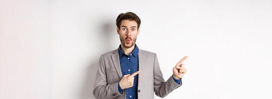 Excited handsome man in suit pointing fingers left at banner and saying wow, look here gesture, showing advertisement, standing on white background photo