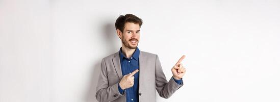 Handsome bearded businessman in grey suit pointing fingers left at logo, inviting check out advertisement, standing on white background photo