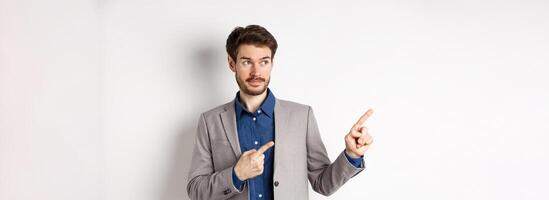 Doubtful male entrepreneur in suit looking and pointing left with hesitant face, standing unsure against white background photo