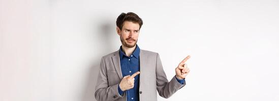 Displeased businessman in suit pointing and looking left with skeptical smirk, dislike banner, disapprove promo offer, standing on white background photo