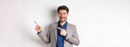 Happy businessman in suit pointing fingers right and smiling excited, showing special deal, standing on white background photo