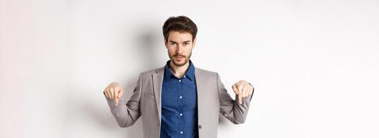 Look here. Handsome bearded man in suit pointing fingers down, looking confident at camera, showing banner, standing on white background photo