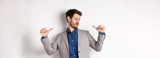 Handsome and confident man in suit pointing at himself, feeling cool and sassy, self-promoting his good appearance, standing on white background photo