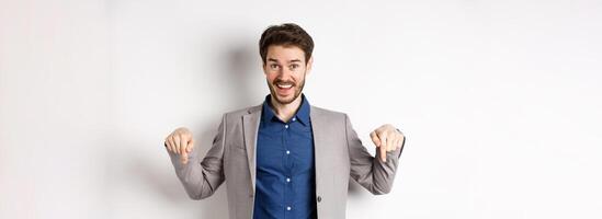 Hey check this out. Happy businessman in suit pointing fingers down and smiling, inviting to event, showing link, standing on white background photo