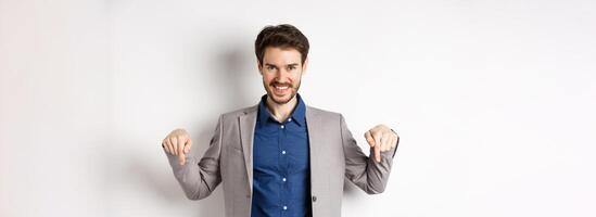 Smiling handsome man in business suit pointing fingers down, showing good deal, look here gesture, standing on white background photo