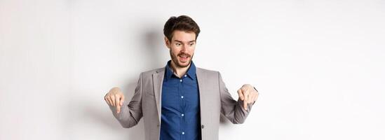 Excited guy in suit smiling and pointing fingers down, looking at special deal with amused face, standing on white background photo