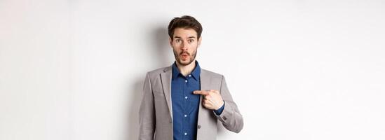 Who me. Surprised man in suit pointing at himself with disbelief, looking confused at camera, standing on white background photo