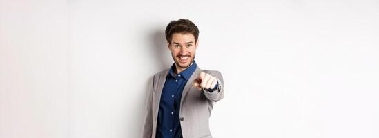 Its you. Smiling handsome manager in suit pointing at camera, choosing person and looking pleased, standing on white background photo
