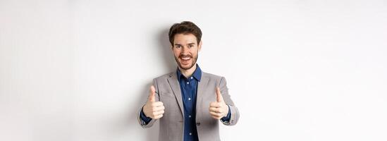 Cheerful and positive young man in business suit showing thumbs up and smiling, like something good, praise good job, standing happy on white background photo
