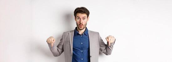 Wow look down. Excited business guy in suit pointing fingers bottom advertisement and look amazed, standing on white background photo
