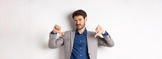 It sucks. Man feeling awkward showing thumbs down and judging something bad, frowning disappointed, standing in suit on white background photo