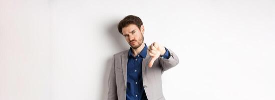 Negative emotions. Disappointed businessman frowning, showing thumb down to disapprove product, dislike something bad, stading on white background photo