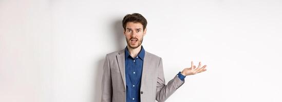 Whats wrong. Confused guy in suit cant understand, looking clueless with hand raised up, staring at camera questioned, standing on white background photo