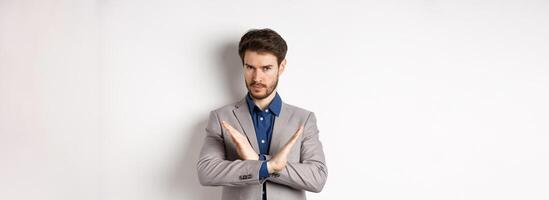 Serious business man making cross and says no, looking determined, disagree and prohibit something bad, tell to stop, standing in suit on white background photo