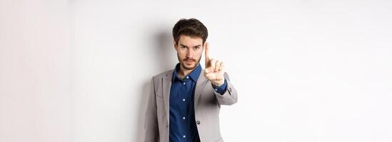 Listen to me. Serious male ceo manager showing one finger, scolding or telling to stop, tell no, hold right there sign, standing on white background in suit photo