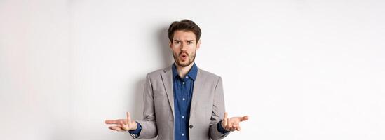 Confused and shocked businessman raising hands up and asking what happened, frowning disappointed, waiting for explanation, standing in suit on white background photo