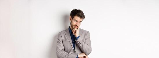 Hesitant businessman in suit making decision, looking pensive at camera with concerned face, having doubts, standing on white background photo