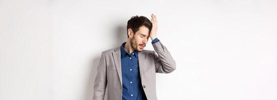 Annoyed businessman in suit facepalm with eyes closed, bothered by someone stupid, pissed-off at work, standing on white background photo