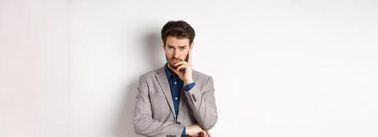 Troubled frowning business man in suit looking at camera pensive, thinking or making decision, standing on white background photo