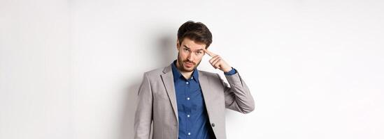 Annoyed male employer scolding personal, pointing finger at head, are you stupid out of your mind gesture, stare at camera from under forehead, standing in suit on white background photo