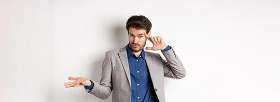 Are you crazy. Young man in suit pointing finger at head to scold strange or stupid mistake, shrugging shoulders confused, standing on white background photo