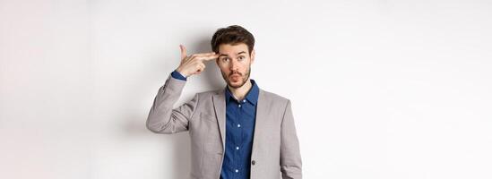 Annoyed guy in business suit shoot himself with hand gun near head, look distressed and tired after work, standing on white background photo