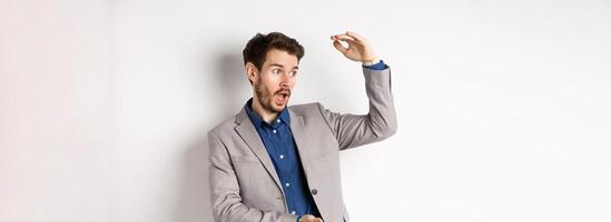 Handsome salesman in suit showing big size, large profit, smiling at camera enthusiastic, standing on white background photo