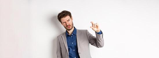 Businessman trying to see something tiny, showing small thing size with fingers, standing in suit on white background photo