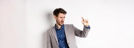 Businessman trying to see something small, showing little thing size with fingers, standing in suit on white background photo