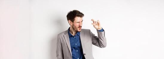 Businessman trying to see something tiny, showing little thing size with fingers, standing in suit on white background photo
