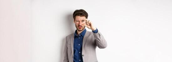 Businessman trying to see something tiny, showing small thing size with fingers, standing in suit on white background photo