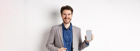 Handsome smiling salesman in suit showing empty smartphone screen, demonstrate an app, standing on white background photo