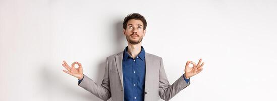 Calm ceo manager in suit relaxing with meditation, looking up and holding hands in zen mudra sign, standing on white background photo