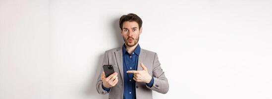 Excited guy in suit say wow, found something online on smartphone, pointing at mobile phone impressed, white background photo