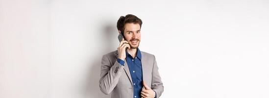 Successful businessman fixing his suit and talking on mobile phone, smiling at camera, standing on white background photo