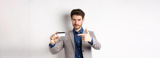 Successful businessman in suit pointing at plastic credit card and smiling, recommending bank, standing on white background photo