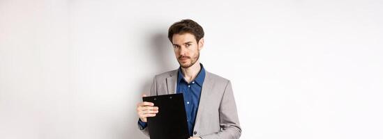 Young male ceo manager holding clipboard with documents and looking at camera, inspecting office, standing on white background photo