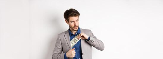 Confident businessman put money in suit and looking aside, standing on white background photo