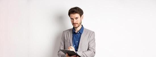 Successful businessman in grey suit taking notes, working in office clothing, holding clipboard and looking at camera, white background photo