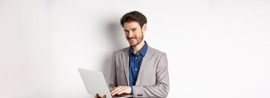 Successful smiling businessman working on laptop and looking happy at camera, standing in grey suit on white background photo