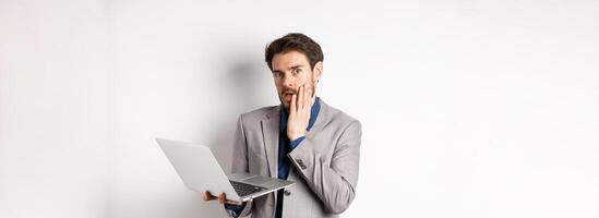 Shocked and troubled businessman have trouble on laptop, looking worried at camera, made mistake at work, standing on white background in suit photo