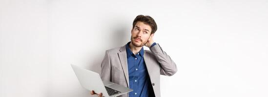 Thoughtful businessman scratch head and look away, making decision while working on laptop, standing against white background photo
