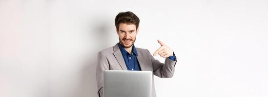 Confident young male entrepreneur working online with laptop, pointing at computer and smiling, standing on white background photo