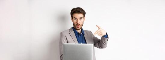 Excited handsome man asking to look here, pointing at laptop screen with amazed face, standing in business suit against white background photo