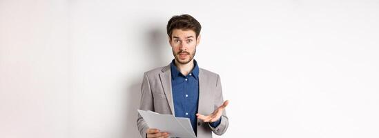 Confused businessman looking at bad document, complaining on deal, pointing at paper frustrated, standing in suit on white background photo