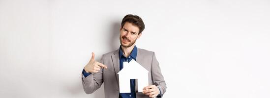 Real estate and insurance concept. Salesman in grey suit showing paper house cutout, selling property, smiling at camera, white background photo