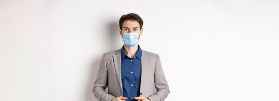 Covid-19 and office workers concept. Confident businessman in medical mask and suit looking at camera, standing on white background photo
