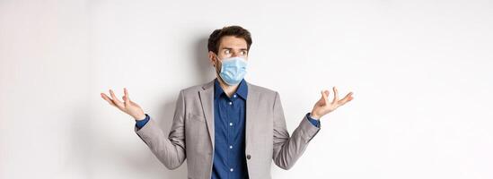 Confused businessman in medical mask shrugging and looking aside, know nothing, cant understand something, standing on white background photo