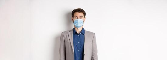 Health, pandemic and business concept. Surprised guy in suit and medical mask raising eyebrows, stare shocked at camera, white background photo